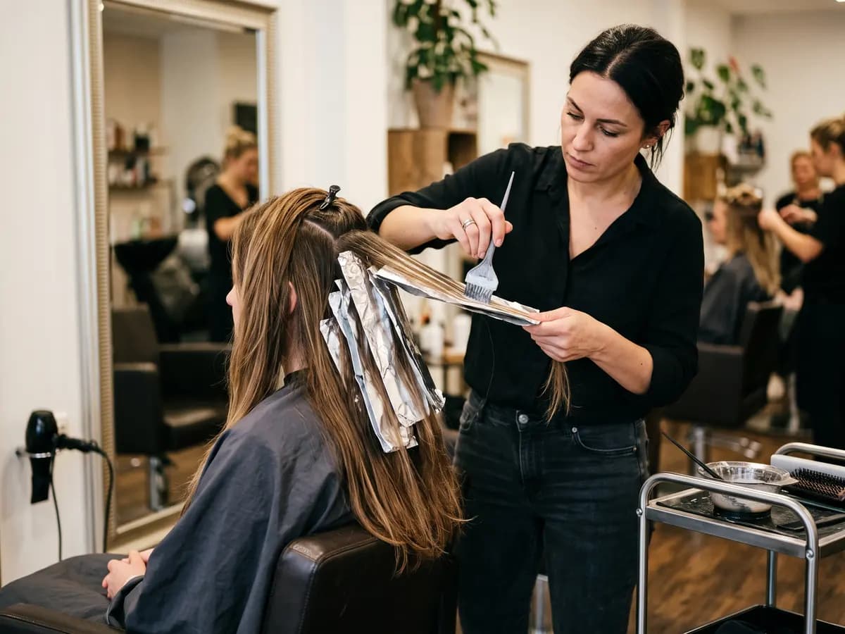 Eine Frau in schwarzer Bluse streicht mit dem Pinsel Farbe auf eine Haarsträhne über Alufolie, die Person im Friseurstuhl trägt einen grauen Umhang